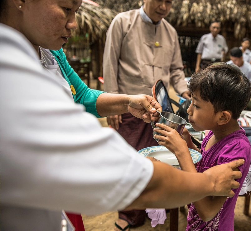 Foto de um grupo de pessoas. Ao centro, um menino moreno é assistido por uma pessoa médica e ao redor outros adultos profissionais de saúde. O ambiente é rural, tropical. 