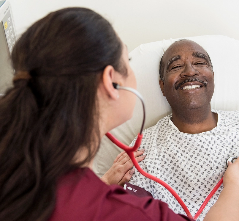 Foto de um homem deitado na maca com sorrisso no rosto sendo examinado por uma pessoa médica usando estetoscópio. 