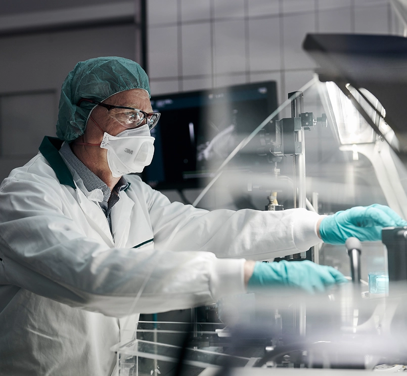 Foto de um homem equipado com jaleco, máscara, óculos de segurança e luvas. Ele mexe em objetos através de um vidro iluminado por luz branca. 