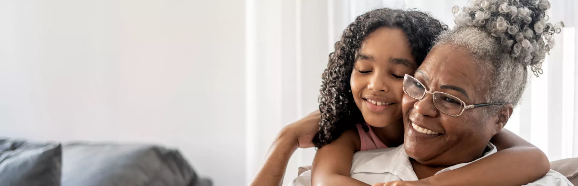 Foto de uma mulher idosa sentada, abraçada por trás por uma jovem criança. A mulher é negra, de cabelos brancos usa óculos e está sorrindo, ergue a mão para apoiar na menina. A criança é uma menina negra, de cabelos longos cacheados e está sorrindo de olhos fechados no abraço. 