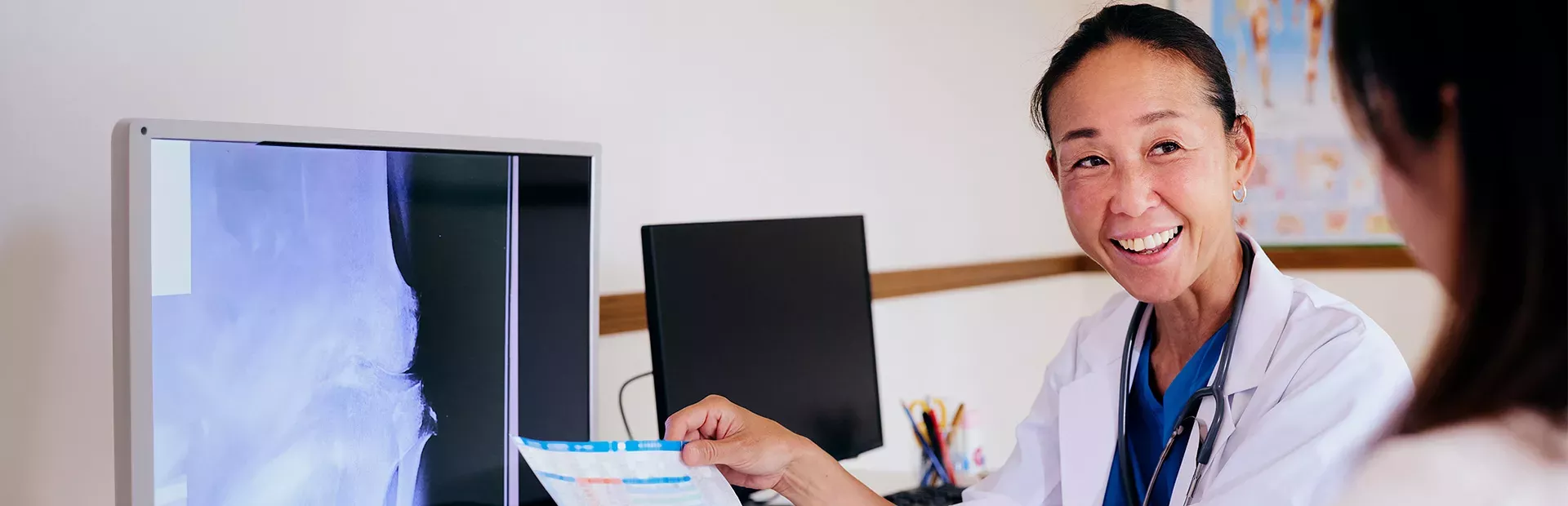 Foto de uma mulher olhando para o lado sorrindo enquanto segura uma folha de papel com informações escritas nessa folha. Ao lado dela há monitores com telas de raio-x.  A mulher é adulta, asiática, de cabelo escuro preso em rabo de cavalo, veste jaleco branco com uniforme hospitalar azul por debaixo. 