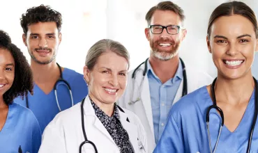 Foto de uma equipe hospitalar de 5 pessoas sorrindo para a câmera. Da direita para esquerda, uma mulher negra de cabelo médio crespo com uniforme azul, um homem pardo de barba e bigode escuros e uniforme azul, uma mulher branca com cabelo preso e jaleco branco, um homem branco de barca marrom usando óculos e jaleco branco, uma mulher de pele média usando uniforme azul.  Todos usam estetoscópio em volta do pescoço.