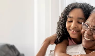 Foto de uma mulher idosa sentada, abraçada por trás por uma jovem criança. A mulher é negra, de cabelos brancos usa óculos e está sorrindo, ergue a mão para apoiar na menina. A criança é uma menina negra, de cabelos longos cacheados e está sorrindo de olhos fechados no abraço. 