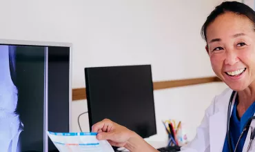 Foto de uma mulher olhando para o lado sorrindo enquanto segura uma folha de papel com informações escritas nessa folha. Ao lado dela há monitores com telas de raio-x.  A mulher é adulta, asiática, de cabelo escuro preso em rabo de cavalo, veste jaleco branco com uniforme hospitalar azul por debaixo. 