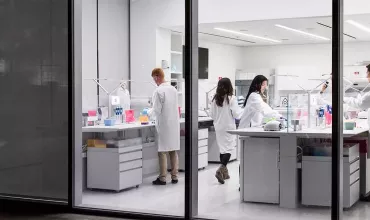 Foto de uma sala de laboratório vista através das janelas altas. O laboratório é iluminado com luz branca, com bancadas, chão e paredes brancas. Nas bancadas estão frascos coloridos e adjacente mulheres e homens de jaleco branco trabalham.