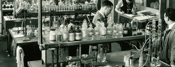 Fotografia antiga do departamento farmacêutico da Sandoz. Foto em preto e branco de uma sala estilo laboratório. Algumas pessoas vestida de jaleco e roupa social estão trabalhando com fracos de vidro dispostos numa mesa de dois níveis, no nível superior existem vários fracos de vidro e na mesa de baixo equipamentos e espaço livre. 
