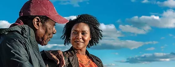 Foto de um homem e uma mulher com fundo de nuvens e céu azul. A mulher é preta com cabelo crespo médio, olha para o homem com sorriso discreto. Ela veste uma jaqueta marrom e blusa laranja. O homem, mais idoso, preto, de cabelo curto e boné vermelho, veste uma jaqueta verde cinzenta e olha para próximo da mulher. 