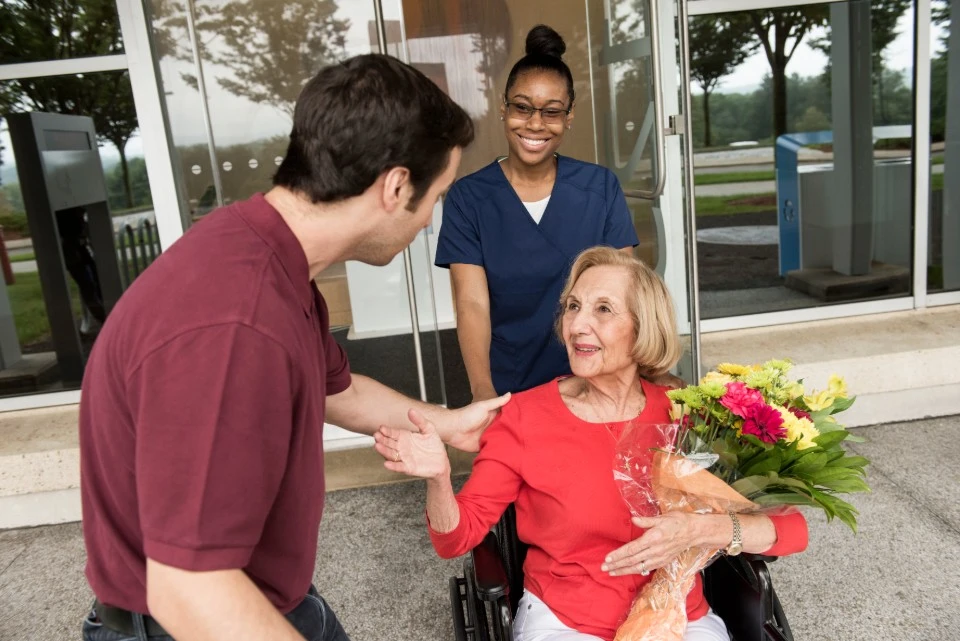 Foto de uma mulher idosa em cadeira de rodas saíndo do prédio com um buquê colorido de flores. Ela é levada por uma enfermeira sorridente com uniforme azul marinho. Ela é recebida por um rapaz jovem de cabelos castanhos. 