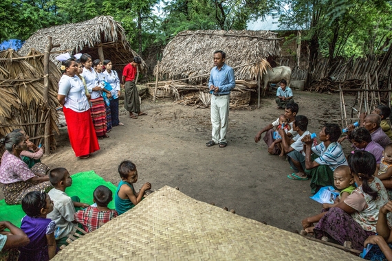 Foto de um local externo rural com um grupo de crianças e adultos atentos a um homem no centro. O ambiente possui estruturas de palha e madeira ao fundo, o chão é de terra, em desfoque atrás estão árvores tropicais. O homem ao centro é negro e veste roupa social azul e bege. As pessoas ao redor são diversas e vestem roupas coloridas como regatas, camisas, shorts e saias. 