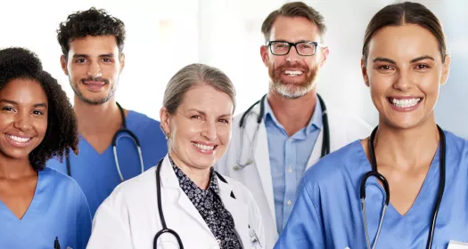 Foto de uma equipe hospitalar de 5 pessoas sorrindo para a câmera. Da direita para esquerda, uma mulher negra de cabelo médio crespo com uniforme azul, um homem pardo de barba e bigode escuros e uniforme azul, uma mulher branca com cabelo preso e jaleco branco, um homem branco de barca marrom usando óculos e jaleco branco, uma mulher de pele média usando uniforme azul.  Todos usam estetoscópio em volta do pescoço.