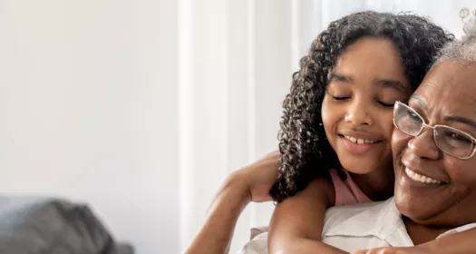 Foto de uma mulher idosa sentada, abraçada por trás por uma jovem criança. A mulher é negra, de cabelos brancos usa óculos e está sorrindo, ergue a mão para apoiar na menina. A criança é uma menina negra, de cabelos longos cacheados e está sorrindo de olhos fechados no abraço. 