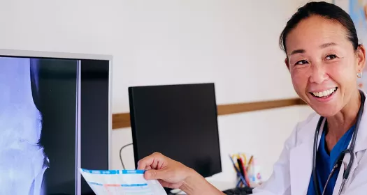 Foto de uma mulher olhando para o lado sorrindo enquanto segura uma folha de papel com informações escritas nessa folha. Ao lado dela há monitores com telas de raio-x.  A mulher é adulta, asiática, de cabelo escuro preso em rabo de cavalo, veste jaleco branco com uniforme hospitalar azul por debaixo. 