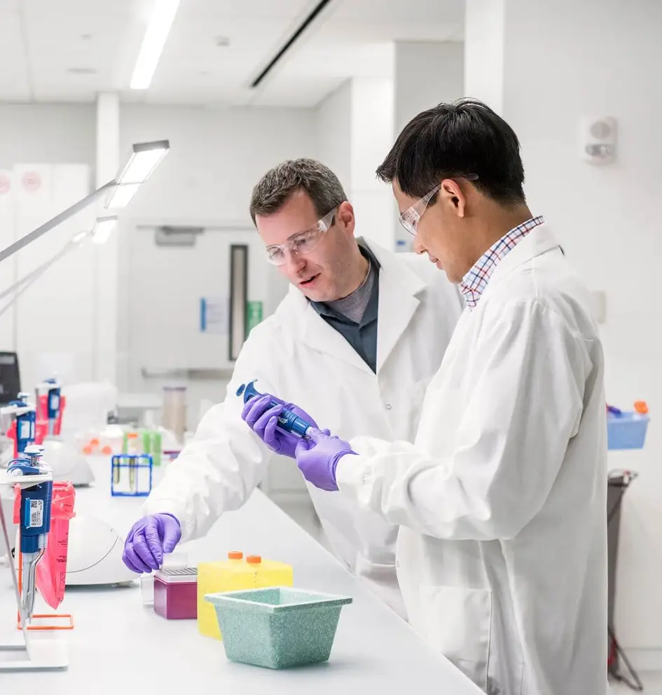 Two scientists in lab coats and safety glasses collaborating on an experiment using a pipette in a laboratory