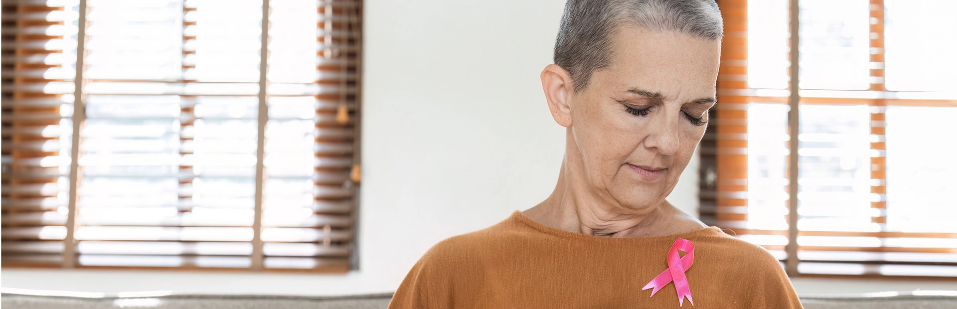 Eine Frau mit kurzgeschorenen Haaren hält die Hände vor der Brust und trägt eine rosa Schleife am Pullover.