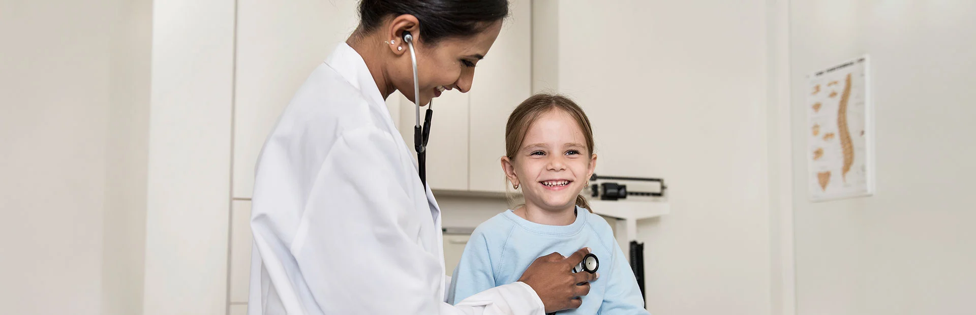 Eine Frau im weißen Kittel horcht ein Mädchen mit einem Stethoskop ab.