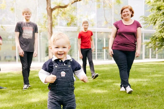 Ein Baby steht lachend im Park und wird von drei weiteren Personen begleitet