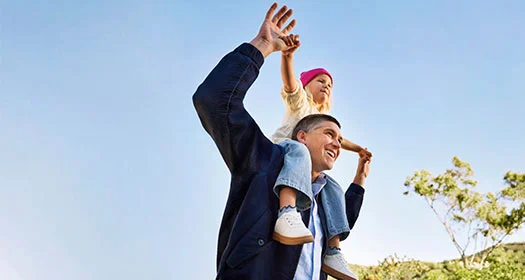 Glücklicher Elternteil mit Kind auf den Schultern unter klarem Himmel.