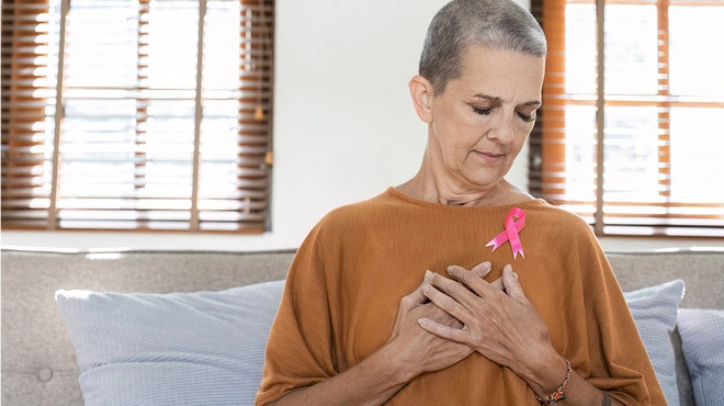Eine Frau mit kurzgeschorenen Haaren hält die Hände vor der Brust und trägt eine rosa Schleife am Pullover.