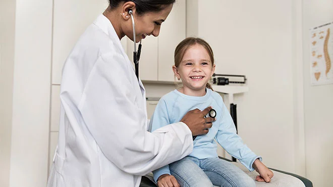 Eine Frau im weißen Kittel horcht ein Mädchen mit einem Stethoskop ab.