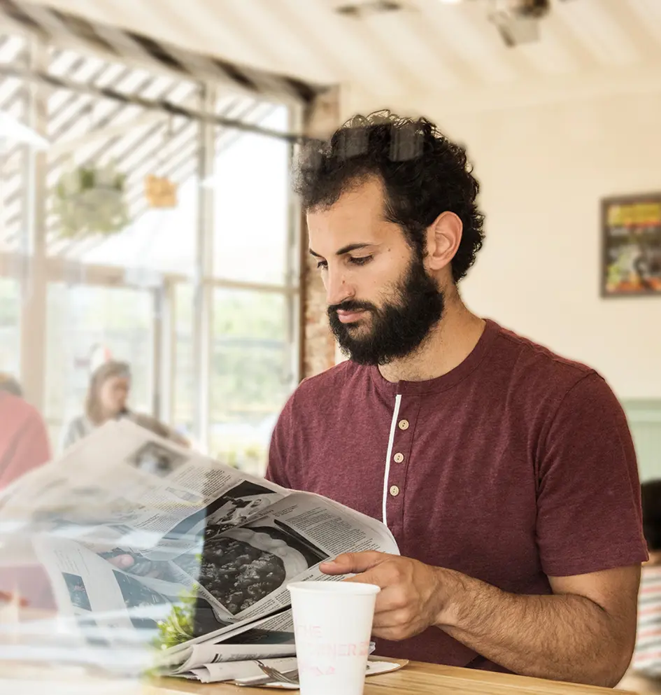 Man reading a newspaper at a bright cafe table.