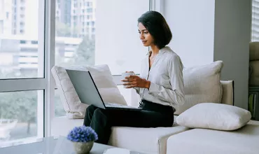 A business woman, seated on a sofa, using a laptop and holding a cup