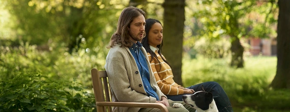 Young couple sitting on a bench in a park with the man looking puzzled.