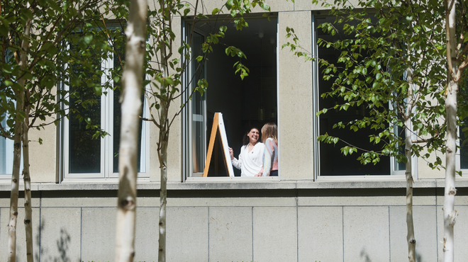 Two people standing by an open window of a modern building behind a row of slender trees.