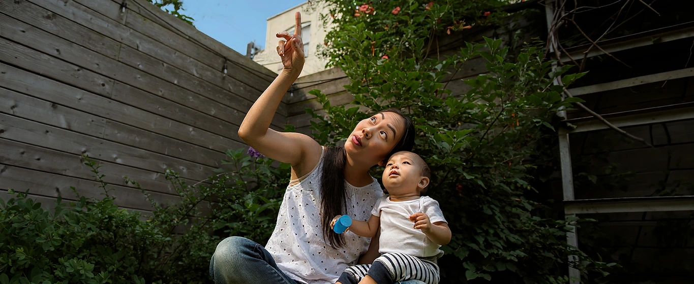 Person holding baby outdoors near gardening plants on a sunny day.
