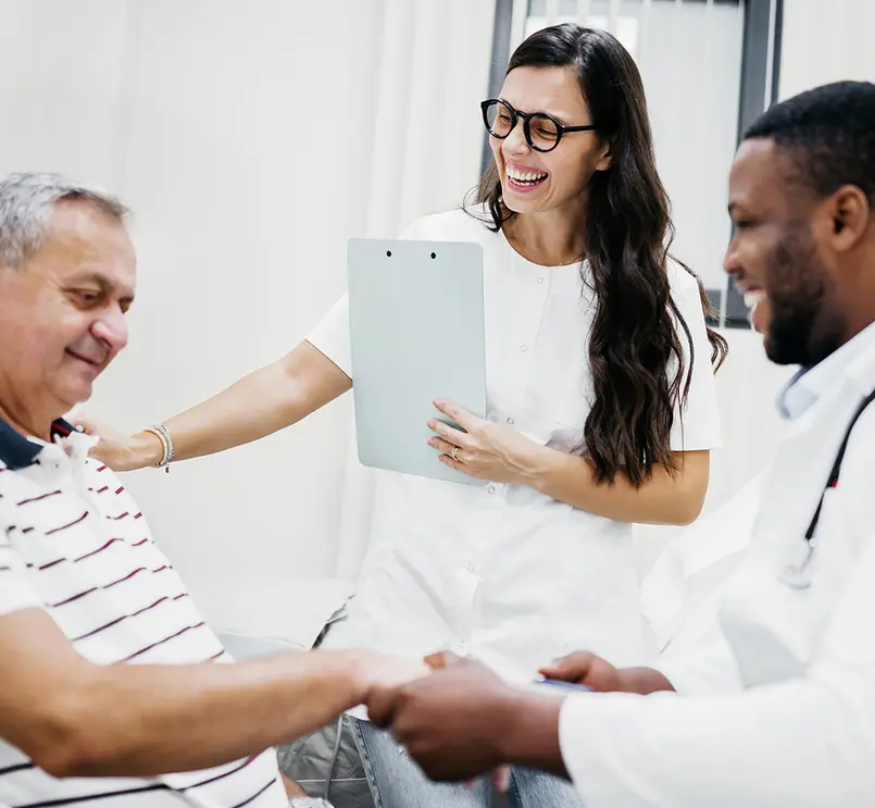 Two smiling healthcare professionals in a medical office, smiling with a patient as they check his pulse.