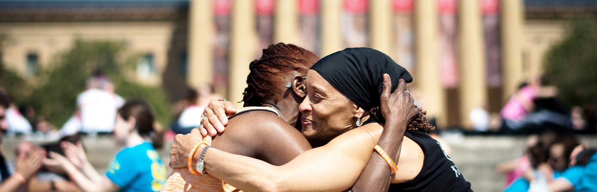 Two smiling women embrace on the steps of a museum, surrounded by event participants in matching shirts. The setting and bright blue sky evokes wellness and community.