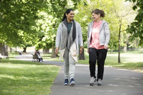 Two women smiling as they walk together through a sunny park, enjoying a friendly moment.  
