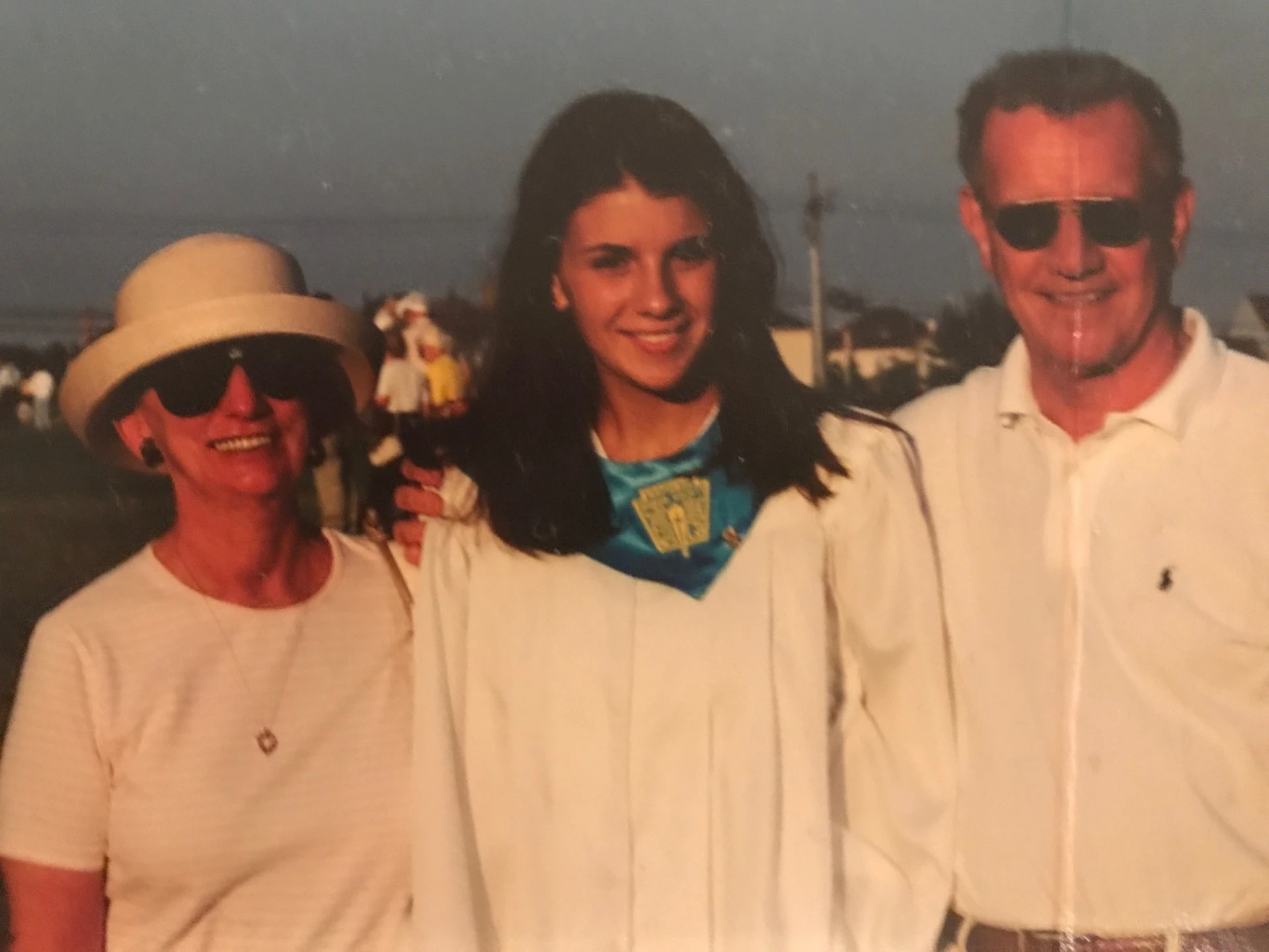A young woman in a graduation gown stands between an elderly couple. They are all smiling, with a crowd and a clear sky in the background.