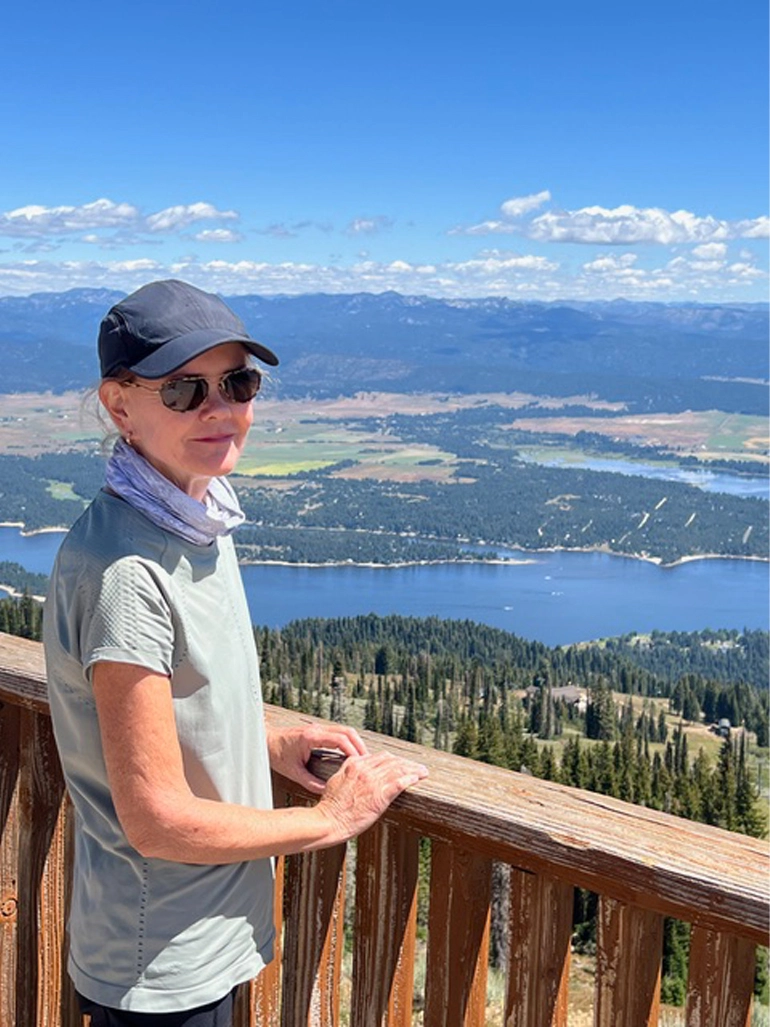Susan shown standing alone on an outdoor balcony overlooking a beautiful landscape of mountains and a lake
