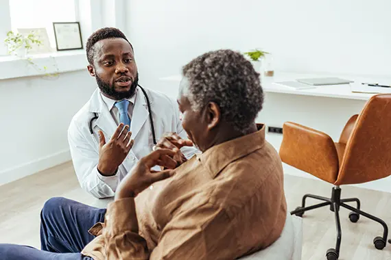 A doctor seated in his office, engaged in an earnest conversation with an older patient. The doctor gestures expressively as he explains information, while the patient listens intently, seated comfortably. The office environment is professional yet inviting.