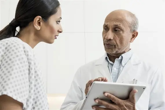A serious yet attentive male doctor holding a tablet, discussing medical information directly with a female patient dressed in a hospital gown. Both are focused on their conversation in a clean, clinical environment.