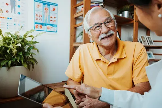 A smiling older man wearing glasses and a yellow polo shirt seated in a medical office, listening attentively to a healthcare professional who is pointing at a tablet screen. Informational posters and plants are visible in the bright background.