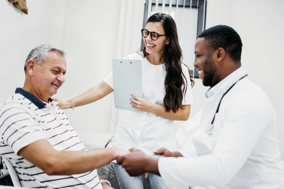 An older male patient seated comfortably, smiling while interacting with two cheerful healthcare professionals. One doctor shakes the patient’s hand warmly, while another stands holding a clipboard, smiling broadly. The atmosphere is positive and supportive.