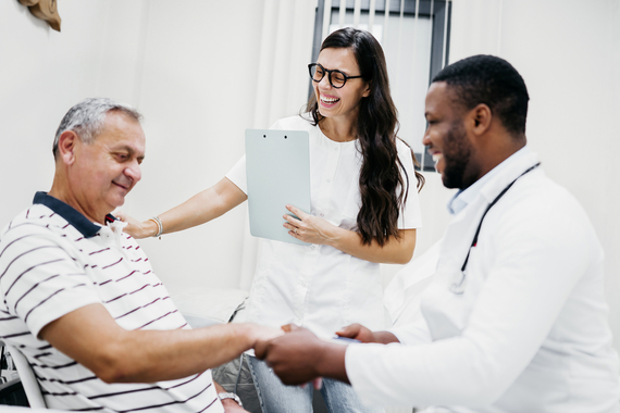 An older male patient seated comfortably, smiling while interacting with two cheerful healthcare professionals. One doctor shakes the patient’s hand warmly, while another stands holding a clipboard, smiling broadly. The atmosphere is positive and supportive.