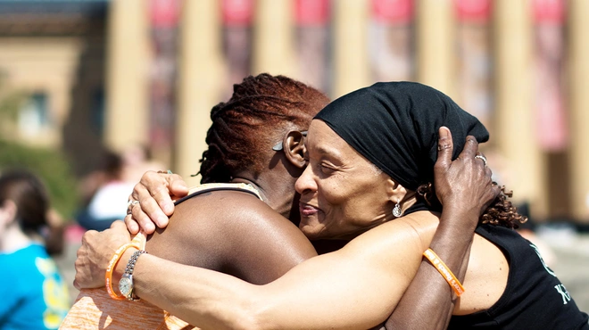 Two smiling women embrace on the steps of a museum, surrounded by event participants in matching shirts. The setting and bright blue sky evokes wellness and community.