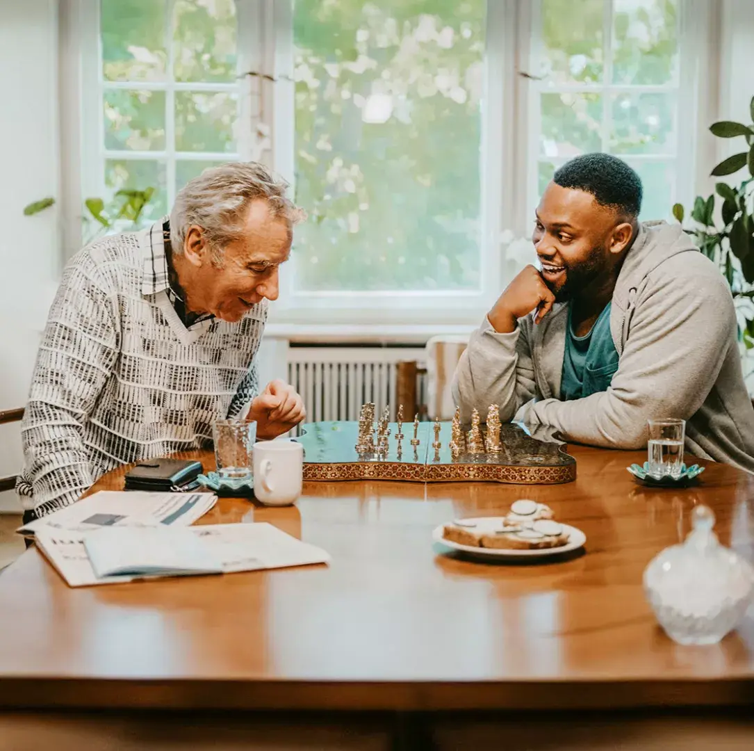 An older man and a younger man playing chess at a table indoors, laughing and engaging in friendly conversation. The room is warmly lit, with plants, newspapers, and refreshments on the table.